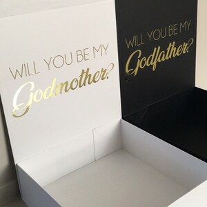 May include: A white box with a black lid. The lid is open and the inside of the box is white. The front of the box has the text "Will you be my Godmother?" in gold lettering. The back of the box has the text "Will you be my Godfather?" in gold lettering.