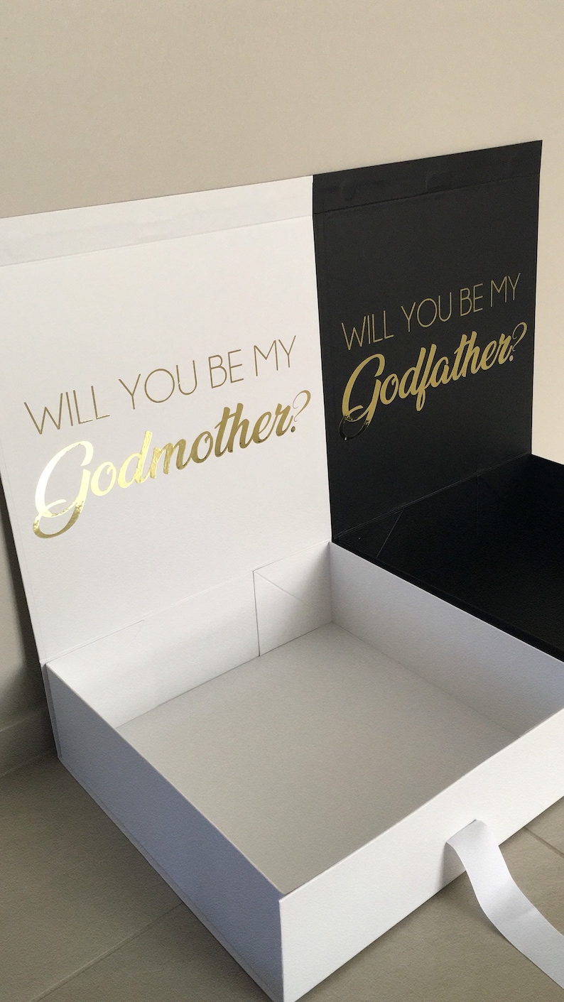 May include: A white box with a black lid. The lid is open and the inside of the box is white. The front of the box has the text "Will you be my Godmother?" in gold lettering. The back of the box has the text "Will you be my Godfather?" in gold lettering.