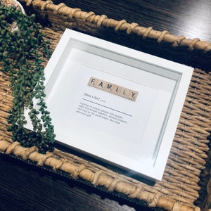 May include: White framed print with the word "FAMILY" spelled out in wooden letter tiles. The definition of the word is below the tiles: "[fam-i-lee] a group of unique people who laugh, play and dream together, always present, not only in the good times; the most precious gift."
