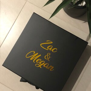 May include: A black gift box with a gold inscription that reads "Zac & Megan".