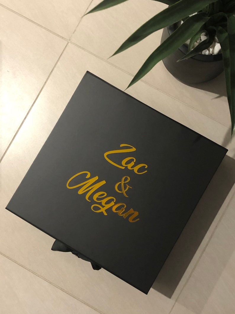 May include: A black gift box with a gold inscription that reads "Zac & Megan".