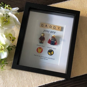 May include: A framed picture with a message that reads "D, A, D, D, Y. you are braver than tougher than You're my favourite superhero! Love Adrian". The picture features two Lego minifigures, one of Iron Man and one of Thor, and their respective logos.