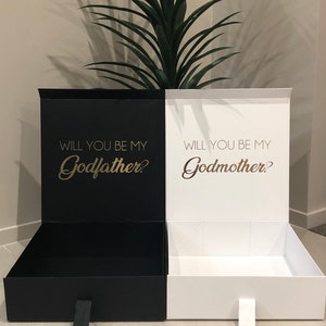 May include: Two black and white gift boxes with lids open. The black box has the text "Will you be my Godfather?" in gold lettering. The white box has the text "Will you be my Godmother?" in gold lettering.