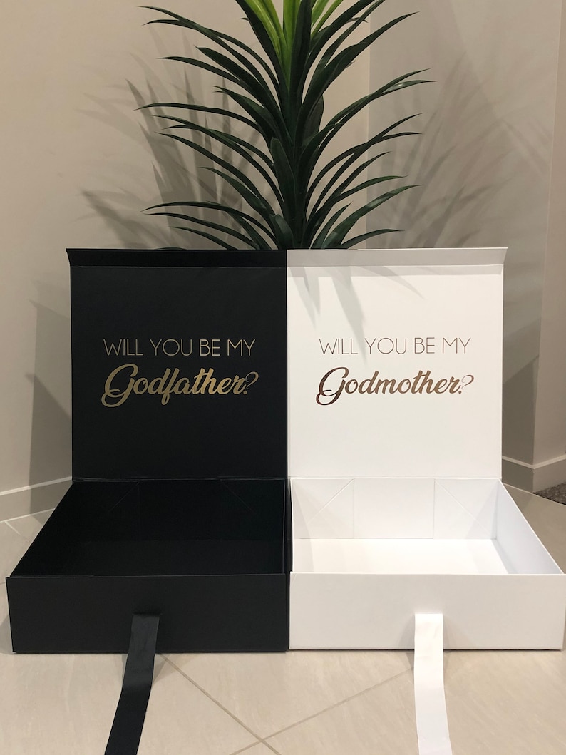 May include: Two black and white gift boxes with lids open. The black box has the text "Will you be my Godfather?" in gold lettering. The white box has the text "Will you be my Godmother?" in gold lettering.