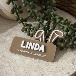 May include: A brown name tag with white bunny ears and the text "Linda Paediatric Audiologist".