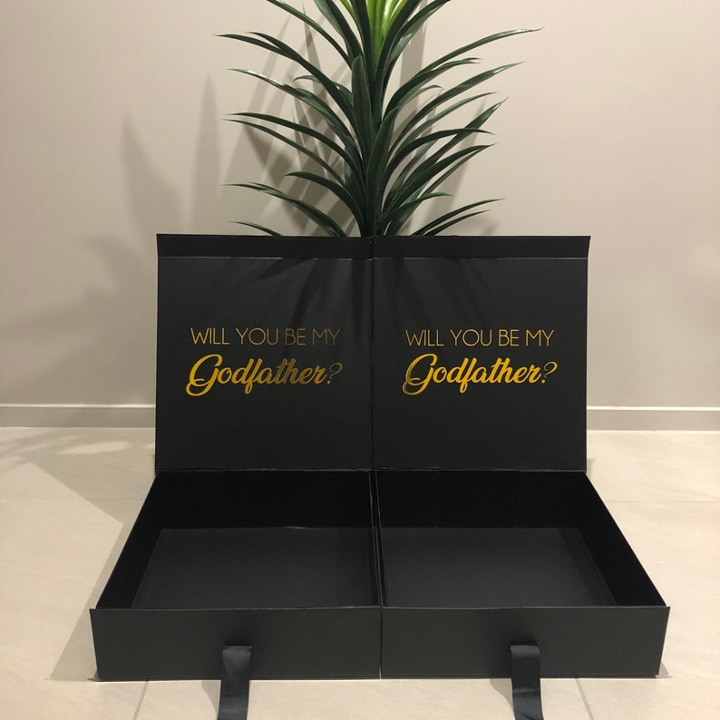 May include: Black gift box with a gold inscription that reads "Will you be my Godfather?" The box is open and has a black interior.
