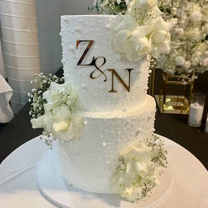 May include: A two-tiered white wedding cake decorated with white pearl sprinkles and white roses. The cake is topped with a gold mirror acrylic topper that says "Z&N".