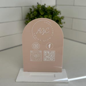 May include: A light pink acrylic sign with a rounded arch top. The sign features the text "Agc Aroma Glow" in a circle with the words "Creative" and "Noilliv" written vertically around the circle. The sign also includes icons for Instagram and Facebook and QR codes for each social media platform.