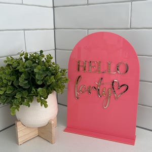 May include: Pink acrylic sign with a gold glitter arch and the text "Hello Forty" with a gold glitter heart.