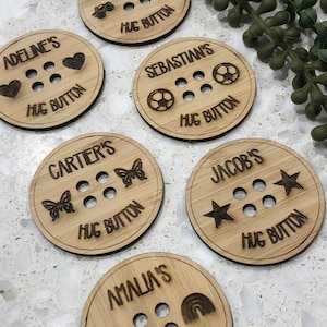 May include: Six wooden round hug buttons with different designs. Each button has a name, a design, and the words "Hug Button". The designs include a heart, a football, butterflies, stars, and a rainbow.