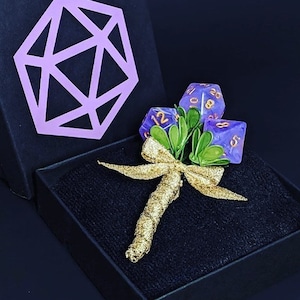 Puede incluir: Un ramo de dados morado y dorado con un lazo dorado y hojas verdes en una caja negra. Los dados tienen números dorados.