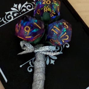 May include: A unique bouquet featuring three iridescent polyhedral dice, each displaying different numbers. The dice are arranged as flower blooms with green fabric leaves. The stem is wrapped in silver glitter, and a silver bow adds a decorative touch.