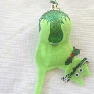 May include: A green felt cat ornament hanging from a green sparkly Christmas bauble. The cat has black eyes and a black tail. The bauble has a gold top and a green string.