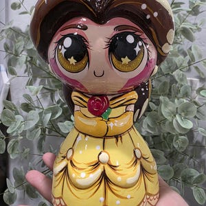 May include: A glossy, hand-painted figurine of a character with large, expressive brown eyes, dark brown hair, and a yellow ball gown. The character holds a small red rose with green leaves at the chest, featuring a smooth, reflective finish.
