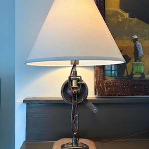 May include: A unique table lamp with a light gray lampshade and a metal base. The base features a wooden platform and a gear-like design. The lamp has a brown power cord and a vintage aesthetic.