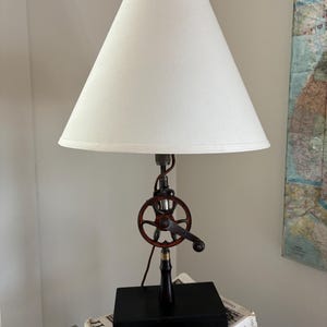 May include: A table lamp featuring a conical off-white fabric lampshade, a central metal hand-crank mechanism with a dark red wheel, and a black rectangular base. The lamp stem is constructed from dark metal components and a black handle.