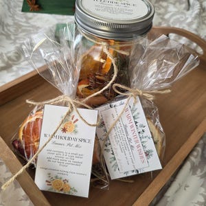 Może przedstawiać: Słoik wypełniony suszonymi plastrami pomarańczy, laskami cynamonu i przyprawami, oznaczony jako "Yuletide Spice". Dwa małe pęczki potpourri związane sznurkiem, każdy z etykietą "Warm Holiday Spice" i "Warm Winter Wishes".