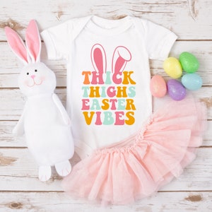 May include: White baby onesie with pink bunny ears and the text "THICK THIGHS EASTER VIBES" in rainbow colors. The onesie is paired with a pink tulle skirt and a white stuffed bunny.