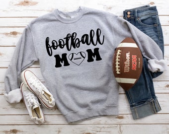 May include: Gray sweatshirt with the text "football mom" in black lettering with a football inside a heart. A brown football and a pair of white sneakers are also in the image.