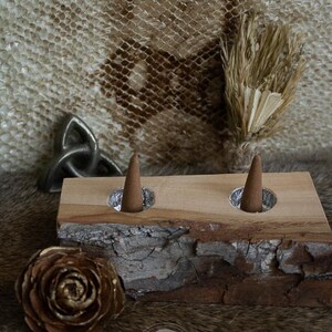 May include: A wooden incense holder with two cone-shaped incense sticks. The holder is made of light-coloured wood with a live edge. A metallic Celtic knot and a gold-tipped pinecone are also visible.