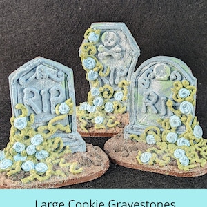 Może przedstawiać: Trzy duże, ozdobne ciasteczka w kształcie nagrobków z napisem "RIP". Ciasteczka są ozdobione zielonym i niebieskim lukrem, który wygląda jak kwiaty. Ciasteczka leżą na brązowej powierzchni.