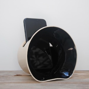May include: A white ceramic phone stand with a black interior. The stand is shaped like a curved horn and has a slot for a phone to be placed in the front.