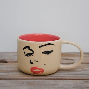 Puede incluir: Una taza de cerámica beige con interior rojo, con un rostro dibujado a mano con cejas negras, pestañas y un diseño de lápiz labial rojo. La taza tiene un asa y está sobre una superficie de madera.