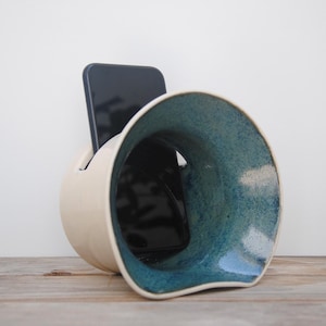 Peut inclure: Support de téléphone en céramique avec un extérieur crème et un intérieur turquoise. Le support a un design incurvé unique avec une grande ouverture. Un smartphone noir est placé à l'intérieur du support, posé sur une surface en bois.