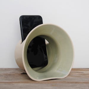 May include: A ceramic phone holder in a light beige and green color scheme. The holder has a unique, curved design with a large opening and a slot to hold a black smartphone. The holder is placed on a wooden surface.