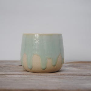 Puede incluir: Una taza de cerámica con un esmalte verde claro que gotea sobre una base beige. La taza es cilíndrica y se encuentra sobre una superficie de madera. El fondo es blanco liso.
