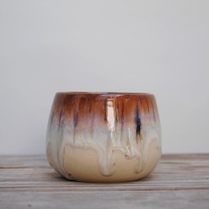 Peut inclure: Un bol en céramique arrondi doté d'un vernis brillant avec un dégradé de couleurs passant d'un brun profond au bord à un blanc crème et un beige clair à la base, marqué par des motifs de coulures verticales sur la surface extérieure.