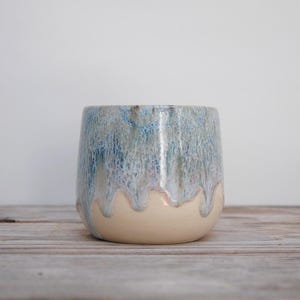 May include: A ceramic tumbler featuring a smooth, matte beige base that transitions into a textured, dripped glaze in shades of blue, grey, and white. The vessel has a rounded, cylindrical shape with a wide opening and a thick, sturdy rim.