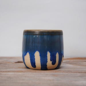 Puede incluir: Un vaso de cerámica hecho a mano con un diseño único. La parte superior es azul oscuro, que pasa a un azul más claro con goteos color crema. El borde es marrón oscuro. El vaso está sobre una superficie de madera.
