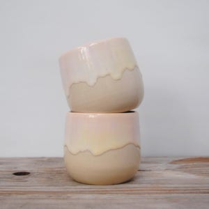 Puede incluir: Dos tazas de cerámica apiladas con un degradado de color. Las tazas transicionan de un rosa suave en la base a un blanco cremoso en la parte superior, con una línea ondulada que separa los colores. Sobre una superficie de madera.