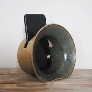 Puede incluir: Un soporte para teléfono de cerámica con un diseño único. El soporte es de color marrón claro con un borde verde oscuro. Un teléfono inteligente negro se coloca dentro del soporte, que se encuentra sobre una superficie de madera.