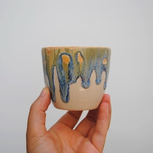 Puede incluir: Una taza de cerámica hecha a mano con una base beige y una sección superior con un esmalte con colores azules, verdes y naranjas goteando. La taza está sostenida en la mano de una persona.