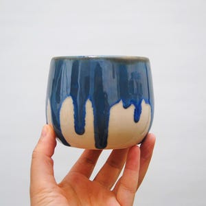 Puede incluir: Una taza de cerámica hecha a mano con forma redondeada. La mitad superior es un esmalte azul oscuro que gotea sobre una base de color crema. La taza se sostiene en la mano de una persona sobre un fondo blanco liso.