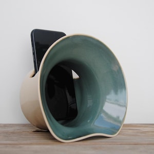 Puede incluir: Un soporte para teléfono de cerámica en una combinación de colores crema y verde azulado. El soporte tiene una forma redondeada con una gran abertura y una ranura para sujetar un teléfono. El interior es verde azulado y el exterior es crema. El teléfono es negro.