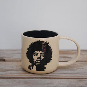 Puede incluir: Taza de cerámica color crema con una silueta negra de una persona con afro. El interior de la taza es negro. La taza tiene un asa redondeada y está sobre una superficie de madera.