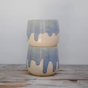 Puede incluir: Dos macetas de cerámica apiladas con un esmalte azul claro que gotea sobre una base crema. Las macetas son cilíndricas con una base redondeada. El fondo es gris claro y las macetas están sobre una superficie de madera. Cada maceta mide unos 10 cm de alto.