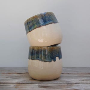 Puede incluir: Dos macetas de cerámica apiladas. Son de color crema con un esmalte azul oscuro y verde alrededor del borde. Las macetas están sobre una superficie de madera. Cada maceta mide unos 10 cm de alto.