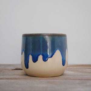 Puede incluir: Un vaso de cerámica con un diseño único. La mitad superior es de color azul oscuro que gotea sobre la base beige. El borde es de color marrón oscuro. El vaso está sobre una superficie de madera.
