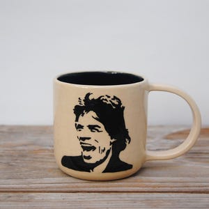 Puede incluir: Una taza de cerámica beige con interior negro y una silueta negra de un rostro. La taza tiene un asa curvada y está sobre una superficie de madera.