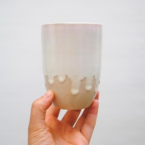 Puede incluir: Una taza de cerámica blanca con un diseño de glaseado goteante. El glaseado es de color marrón claro y gotea por los lados de la taza.
