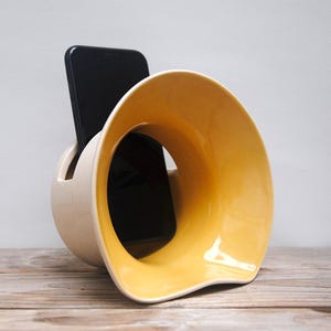 Puede incluir: Un soporte de teléfono de cerámica con un diseño amarillo y blanco. El soporte tiene una forma curva y un agujero en el centro para sujetar un teléfono.