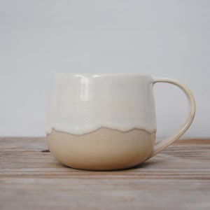 Puede incluir: Taza de cerámica hecha a mano con forma redondeada y un asa grande. La taza es de dos tonos, con un esmalte blanco crema en la mitad superior y un esmalte beige en la mitad inferior. La taza está sobre una superficie de madera.