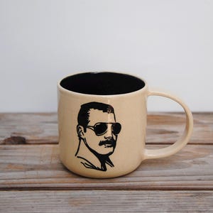 Puede incluir: Una taza de cerámica beige con interior negro y asa. La taza presenta una ilustración en blanco y negro de un hombre con gafas de sol y bigote. La taza está sobre una superficie de madera.