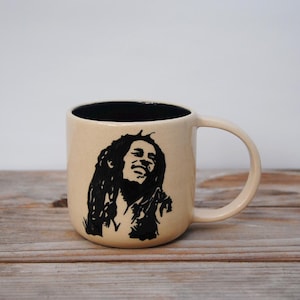Puede incluir: Una taza de cerámica beige con interior negro y asa. La taza presenta una silueta negra de una persona con pelo largo. La taza está sobre una superficie de madera.