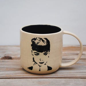 Puede incluir: Una taza de cerámica blanca con una silueta negra de una mujer con el pelo corto. La taza tiene un interior negro y un asa.