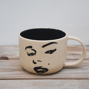 Puede incluir: Una taza de cerámica beige con interior negro y asa. La taza presenta un dibujo lineal negro de una cara en la parte delantera. La cara tiene los ojos cerrados, una marca de belleza y labios llenos. La taza está sobre una superficie de madera.